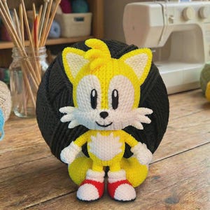 Może przedstawiać: Ręcznie robiona szydełkowa lalka Tails z serii Sonic the Hedgehog. Lalka ma kolory ż&oacute;łty, biały i czerwony, z uśmiechniętą twarzą i dwoma ogonami. Lalka ma około 15 cm wysokości i jest ustawiona na ciemnoszarej kuli przędzy.