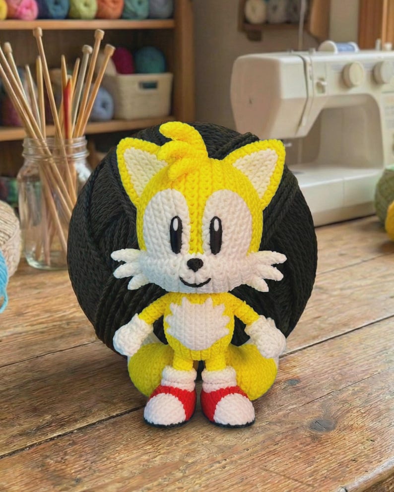 Może przedstawiać: Ręcznie robiona szydełkowa lalka Tails z serii Sonic the Hedgehog. Lalka ma kolory ż&oacute;łty, biały i czerwony, z uśmiechniętą twarzą i dwoma ogonami. Lalka ma około 15 cm wysokości i jest ustawiona na ciemnoszarej kuli przędzy.