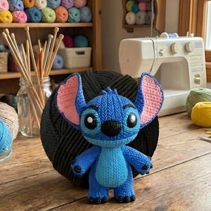 Może przedstawiać: Niebieska dziergana lalka Stitch z r&oacute;żowymi uszami wewnętrznymi i czarnymi oczami, przed czarną kulą z przędzy. Lalka stoi na drewnianej powierzchni, z akcesoriami do robienia na drutach i przędzą w tle.
