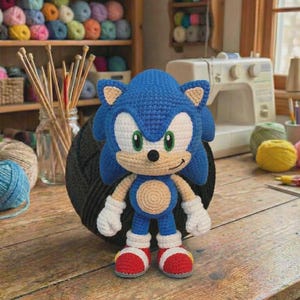 Może przedstawiać: Szydełkowa lalka Sonic the Hedgehog w kolorze niebieskim, białym i czerwonym. Lalka ma uśmiechniętą twarz z zielonymi oczami, białymi rękawiczkami i czerwonymi butami. Ustawiona na drewnianym stole, z przędzą i akcesoriami do szycia w tle.