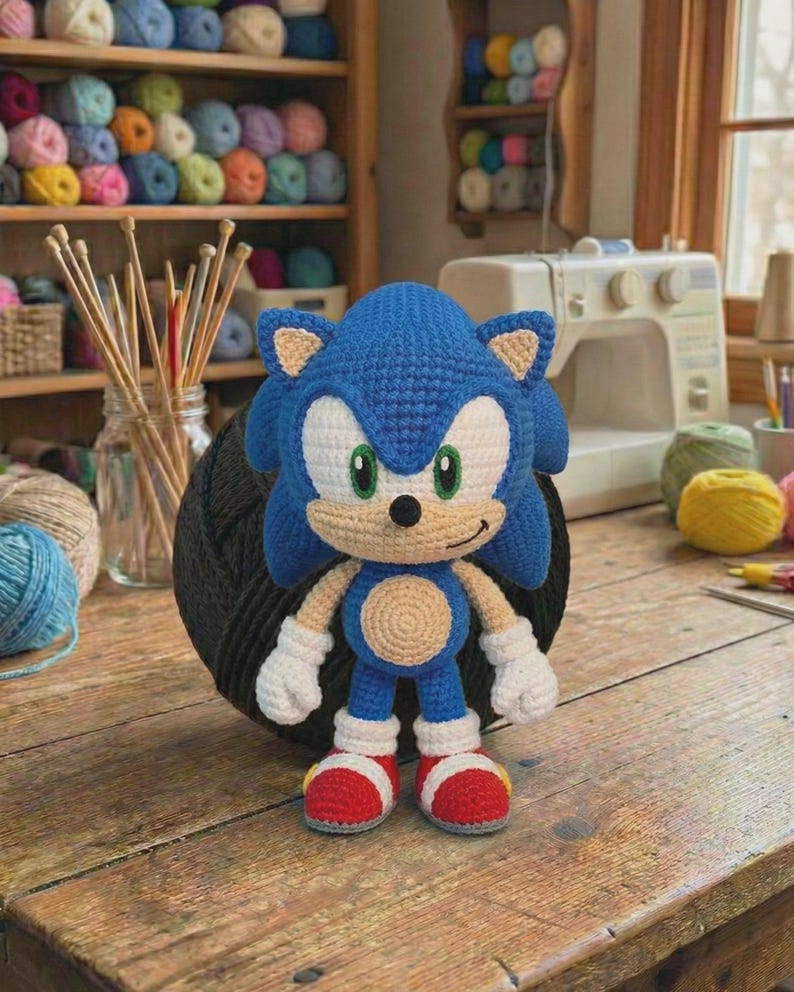 Może przedstawiać: Szydełkowa lalka Sonic the Hedgehog w kolorze niebieskim, białym i czerwonym. Lalka ma uśmiechniętą twarz z zielonymi oczami, białymi rękawiczkami i czerwonymi butami. Ustawiona na drewnianym stole, z przędzą i akcesoriami do szycia w tle.