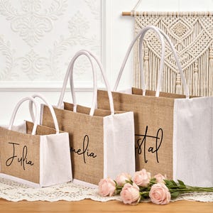 May include: Three burlap tote bags with white accents and handles. Each bag has a name printed in black script: Julia, Amelia, and Crystal. Pink roses are arranged in front of the bags.