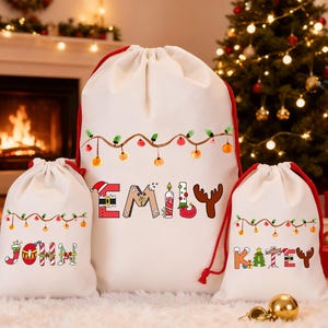 May include: Three white Christmas gift bags with red drawstrings. The largest bag has the name "EMILY" in colourful letters, and the smaller bags say "JOHN" and "KATEY". Each bag has a string of lights design.