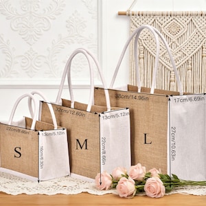 May include: Three jute tote bags in sizes S, M, and L, with white handles and bases. The bags are labeled with their sizes and measurements: S is 8.66in x 6.30in, M is 12.60in x 8.66in, and L is 14.96in x 10.63in. Pink roses are in front.