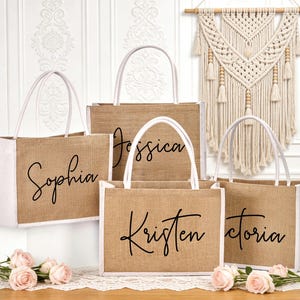 May include: Five burlap tote bags with white handles and trim. Each bag has a different name in black script: Sophia, Jessica, Kristen, and Victoria. The bags are arranged on a table with pink roses.