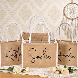 May include: Five burlap tote bags with white accents and handles. Each bag has a name in black script: Kristen, Sophia, and Amelia are visible. The bags are arranged on a lace tablecloth with roses.