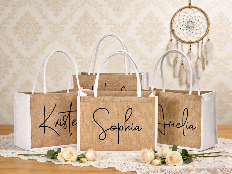May include: Five burlap tote bags with white accents and handles. Each bag has a name in black script: Kristen, Sophia, and Amelia are visible. The bags are arranged on a lace tablecloth with roses.