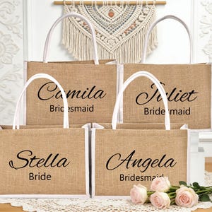 May include: Four burlap tote bags with white handles and trim. Each bag has a name printed in black script: Camila, Juliet, Stella, and Angela. The text "Bridesmaid" or "Bride" is printed below the names. A bouquet of pink roses rests in front.
