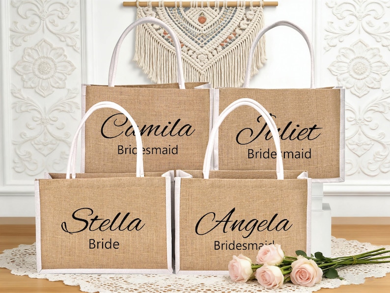 May include: Four burlap tote bags with white handles and trim. Each bag has a name printed in black script: Camila, Juliet, Stella, and Angela. The text "Bridesmaid" or "Bride" is printed below the names. A bouquet of pink roses rests in front.