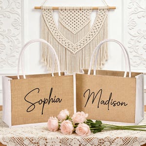 May include: Two jute tote bags with white accents and handles, each personalized with a name in black script: "Sophia" and "Madison." The bags are displayed with pink roses on a lace tablecloth.