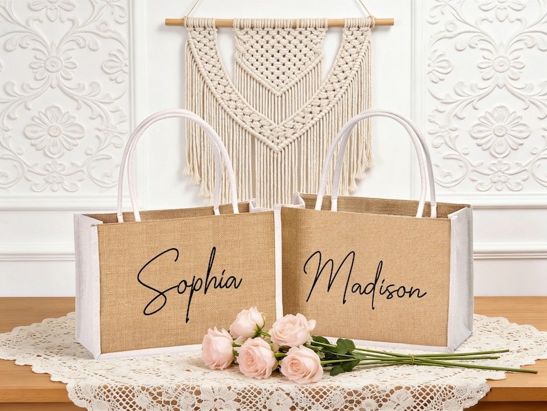 May include: Two jute tote bags with white accents and handles, each personalized with a name in black script: "Sophia" and "Madison." The bags are displayed with pink roses on a lace tablecloth.