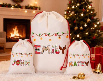 May include: Three white Christmas gift bags with red drawstrings. The largest bag has the name "EMILY" in festive letters, and the smaller bags say "JOHN" and "KATEY". Each bag is decorated with a string of lights.