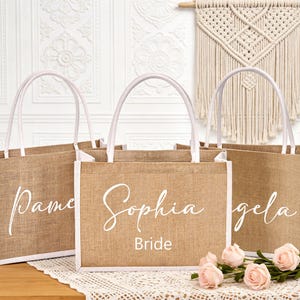May include: Three burlap tote bags with white accents and handles. Each bag has a name in cursive script: Pamela, Sophia, and Angela. The middle bag also says "Bride" below the name. The bags are on a wooden surface with pink roses.