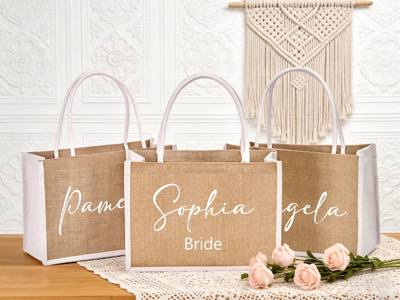 May include: Three burlap tote bags with white accents and handles. Each bag has a name in cursive script: Pamela, Sophia, and Angela. The middle bag also says "Bride" below the name. The bags are on a wooden surface with pink roses.