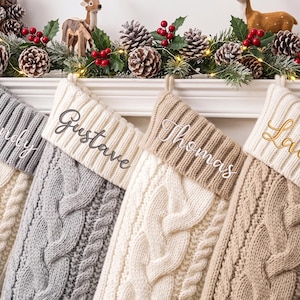 Könnte beinhalten: Vier Weihnachtsstrümpfe aus Zopfstrick in Creme, Hellgrau und Beige. Jeder Strumpf hat ein geripptes Bündchen, das mit den Namen Wendy, Gustave, Thomas und Laurence bestickt ist. Die Strümpfe hängen in einer Reihe an einem weißen Kaminsims.