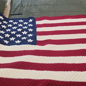 May include: A crocheted blanket featuring the design of the American flag. The blanket has a blue square with white star shapes in the upper left corner, and red and white stripes across the rest of the blanket.