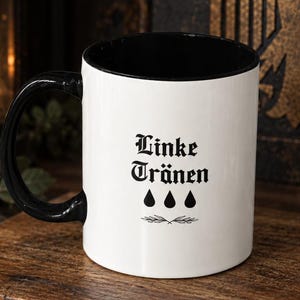 Könnte beinhalten: Eine weiße Keramiktasse mit schwarzem Innenraum und schwarzem Henkel. Auf der Außenseite befindet sich der schwarze Schriftzug Linke Tränen in gotischer Schrift, über drei schwarzen Tränensymbolen und einer kleinen dekorativen Zweiggrafik.