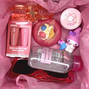 Könnte beinhalten: Eine Geschenkbox mit einer transparenten orangefarbenen Haarklammer, einem Vitamasques Lippenbalsam-Duo, einer rosa Badebombe in Donut-Form mit Creme-Topping, einem Bath & Body Works Tiegel, einer My Melody Figur, Garnier Mizellenwasser und einer roten Sonnenbrille.