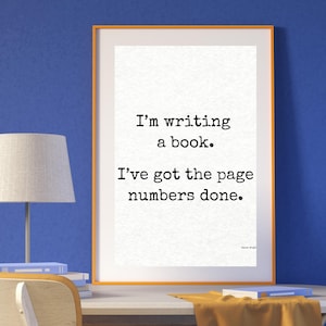 May include: A framed print with the text "I'm writing a book. I've got the page numbers done." The print is in an orange frame, displayed on a white desk with a yellow chair and a lamp.