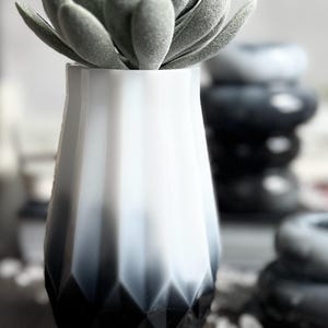 May include: A decorative vase with a geometric design, ombre effect, and a faux succulent. The vase transitions from white to black, with a faceted base. The succulent has soft, gray-green leaves, adding a natural touch to the decor.