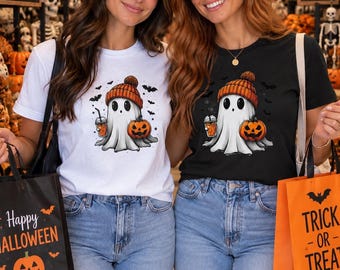 May include: A white t-shirt and a black t-shirt, each featuring a graphic of a ghost wearing an orange knit hat, holding a pumpkin and a drink, surrounded by bats. The shirts are paired with blue jeans and black or orange Halloween tote bags.