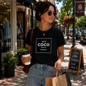 May include: A black short-sleeve t-shirt featuring a white square graphic with the text N&deg;7 COCO MADE IN PARIS printed in the center. The shirt is paired with light blue denim jeans and worn with a gold necklace and round sunglasses.