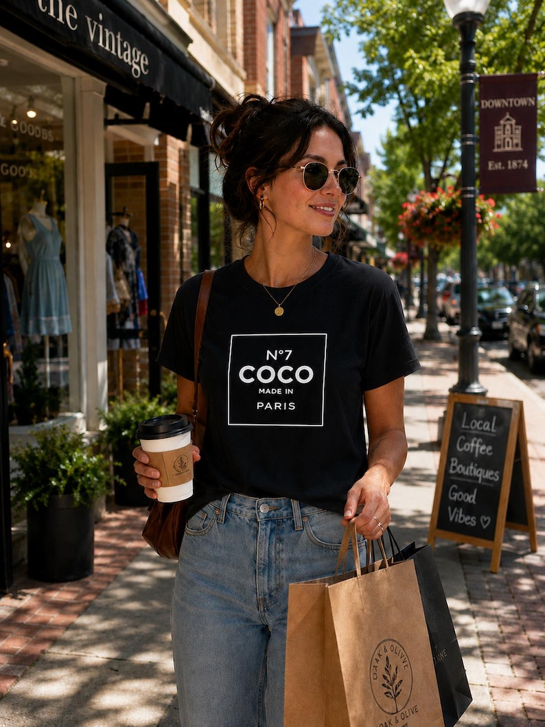 May include: A black short-sleeve t-shirt featuring a white square graphic with the text N&deg;7 COCO MADE IN PARIS printed in the center. The shirt is paired with light blue denim jeans and worn with a gold necklace and round sunglasses.