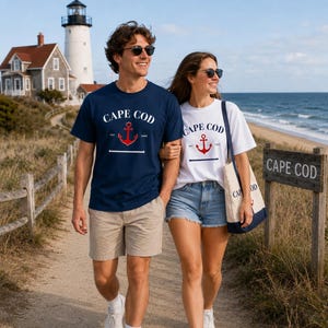 May include: A couple walks along a path near the beach, both wearing sunglasses and t-shirts with a "Cape Cod" anchor design. The man wears a navy blue shirt and khaki shorts, while the woman wears a white shirt and denim shorts. A lighthouse and beach are in the background.