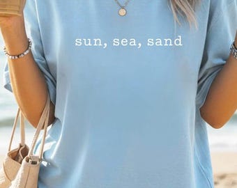 Könnte beinhalten: Hellblaues T-Shirt mit dem weißen Schriftzug "sun, sea, sand". Die Person trägt eine goldene Halskette und hält ein Getränk sowie eine beigefarbene Tragetasche. Der Strand ist im Hintergrund.