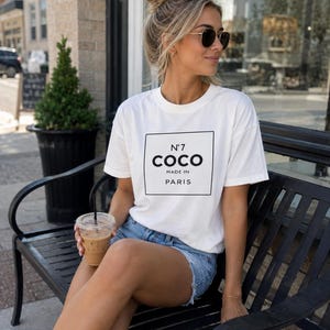 May include: A white short-sleeved t-shirt featuring a black rectangular graphic on the chest with the text N&deg;7 COCO MADE IN PARIS in black lettering, paired with light blue denim shorts and white sneakers.