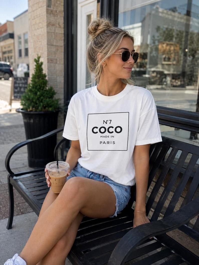 May include: A white short-sleeved t-shirt featuring a black rectangular graphic on the chest with the text N&deg;7 COCO MADE IN PARIS in black lettering, paired with light blue denim shorts and white sneakers.