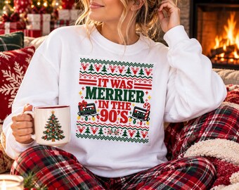 May include: A white long-sleeve sweatshirt featuring a graphic print with the text "IT WAS MERRIER IN THE 90'S" surrounded by red and green holiday patterns, cassette tapes, and string lights, paired with red and black plaid pajama pants.