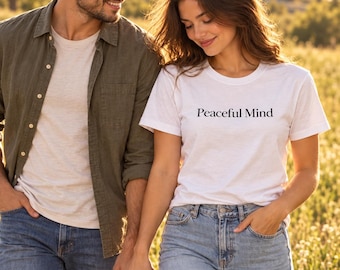 May include: A white crew neck t-shirt featuring the text Peaceful Mind printed in black on the chest. The shirt is paired with light blue denim jeans. The design is simple, focusing on the clear black lettering against the white fabric.