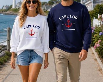 Könnte beinhalten: Ein weißes Sweatshirt und ein marineblaues Sweatshirt, beide mit dem Text CAPE COD in geschwungener Schrift über einer roten Ankergrafik, mit den Buchstaben EST und 1602 auf beiden Seiten des Ankers und einer horizontalen Seilgrafik darunter.