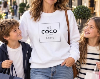 May include: A white long-sleeve crewneck sweatshirt featuring a black square graphic on the chest. Inside the square, the text reads N°7 COCO MADE IN PARIS in black lettering. The sweatshirt has a ribbed neckline, cuffs, and hem.