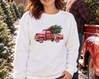 May include: A white long-sleeve crewneck sweatshirt featuring a graphic print of a red vintage pickup truck carrying a decorated Christmas tree in its bed. The design includes snowy details and a festive aesthetic on the front of the garment.