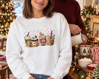May include: A white crewneck sweatshirt featuring a graphic print of four festive holiday beverages in cups, including coffee and cocoa with whipped cream, red straws, and small decorative Christmas tree and candy cane accents.