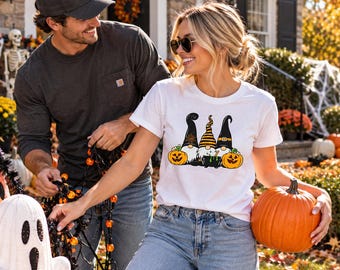 May include: A white short-sleeved t-shirt featuring a graphic print of three gnomes wearing black and orange hats, standing beside two orange jack-o'-lanterns and a black cauldron filled with a green liquid, set against a festive outdoor background.