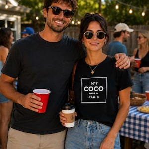May include: A black t-shirt featuring a white square graphic with the text N&deg;7 COCO MADE IN PARIS. The shirt has a crew neckline and short sleeves, paired with light blue denim jeans. The design is centered on the chest in a clean, bold font.