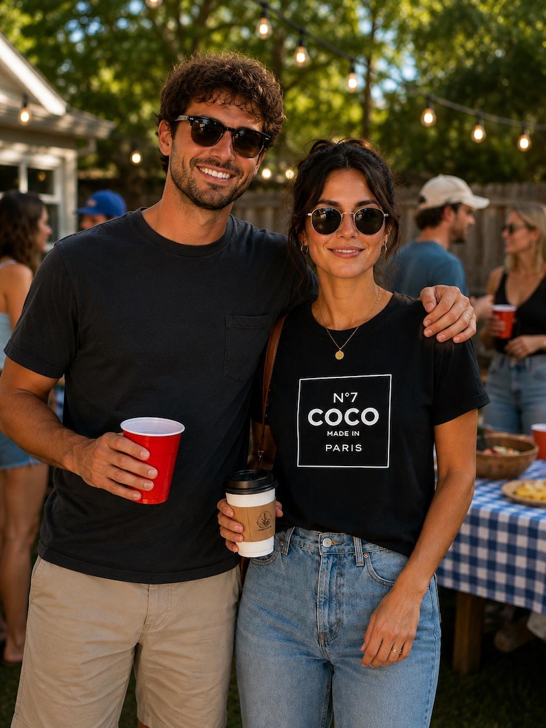 May include: A black t-shirt featuring a white square graphic with the text N&deg;7 COCO MADE IN PARIS. The shirt has a crew neckline and short sleeves, paired with light blue denim jeans. The design is centered on the chest in a clean, bold font.