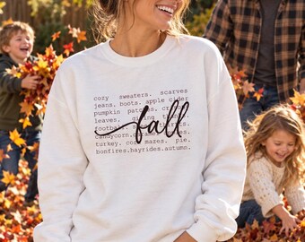 May include: A white crewneck sweatshirt featuring black text listing autumn-themed words like cozy sweaters, scarves, jeans, boots, apple cider, pumpkin patches, crisp leaves, football, candy corn, caramel apples, turkey, corn mazes, pie, and bonfires.