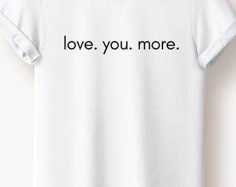 Könnte beinhalten: Weißes T-Shirt mit dem Aufdruck "love. you. more." in Schwarz. Das Shirt hat kurze Ärmel und einen Rundhalsausschnitt. Das Kleidungsstück hängt an einem Holzbügel.