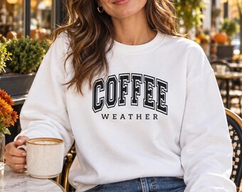 May include: A white crewneck sweatshirt featuring the black text COFFEE WEATHER in a collegiate-style font on the chest. The garment has long sleeves and a ribbed neckline, cuffs, and hem, providing a clean and simple look for casual wear.