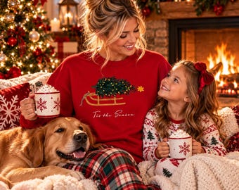 May include: A red crewneck sweatshirt featuring a graphic of a wooden sled carrying a decorated Christmas tree and the text 'Tis the Season' in white script. The sweatshirt is worn with red, green, and white plaid pajama pants.