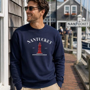 K&ouml;nnte beinhalten: Marineblaues Sweatshirt mit dem Wort "Nantucket" in wei&szlig;em Bogen &uuml;ber einer roten Leuchtturm-Grafik. Das Sweatshirt hat auch den Text "EST 1659" darunter. Die Person tr&auml;gt eine Sonnenbrille und eine khakifarbene Hose.
