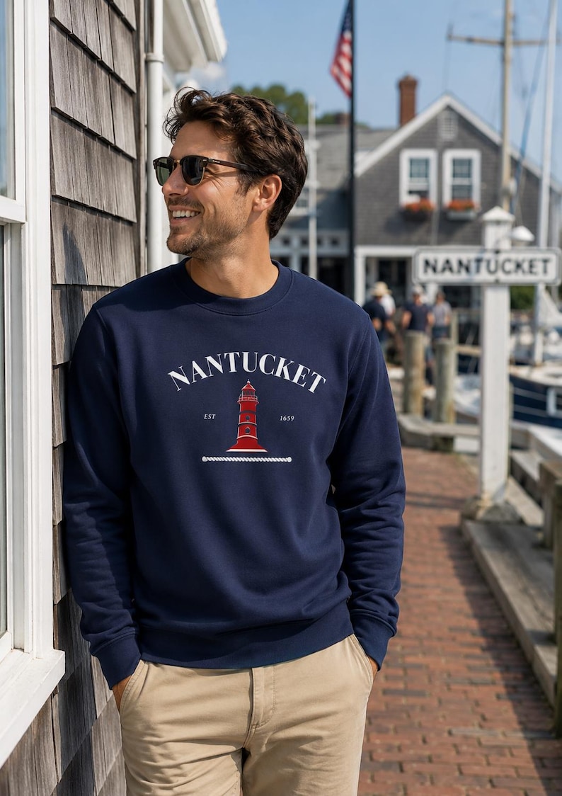 K&ouml;nnte beinhalten: Marineblaues Sweatshirt mit dem Wort "Nantucket" in wei&szlig;em Bogen &uuml;ber einer roten Leuchtturm-Grafik. Das Sweatshirt hat auch den Text "EST 1659" darunter. Die Person tr&auml;gt eine Sonnenbrille und eine khakifarbene Hose.