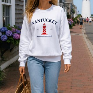 K&ouml;nnte beinhalten: Ein wei&szlig;es Sweatshirt mit dem Wort "Nantucket" in Marineblau, einer roten Leuchtturm-Grafik und dem Jahr "1659". Die Person tr&auml;gt eine hellblaue Jeans, wei&szlig;e Turnschuhe und eine geflochtene Tragetasche.