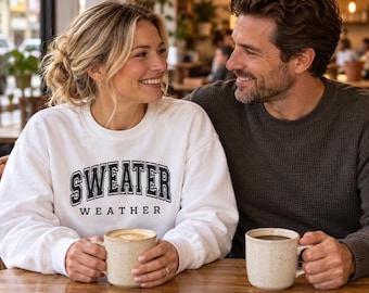 May include: A white crewneck sweatshirt featuring the black text "SWEATER WEATHER" in a collegiate-style font. The garment has long sleeves and a ribbed neckline, cuffs, and hem, providing a relaxed fit for casual wear in cooler temperatures.