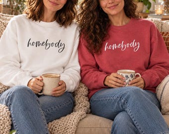 May include: Two crewneck sweatshirts, one white and one red, each featuring the word homebody printed in a cursive script across the chest. The sweatshirts are paired with blue denim jeans and are worn while holding ceramic mugs.