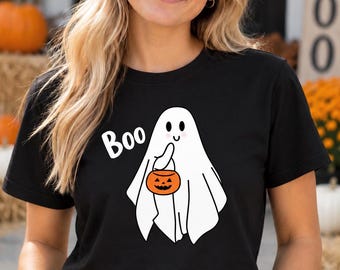 May include: A black short-sleeved t-shirt featuring a white graphic of a ghost holding a small orange jack-o'-lantern bucket. The word Boo is printed in white text next to the ghost, which has simple black eyes and pink cheeks.